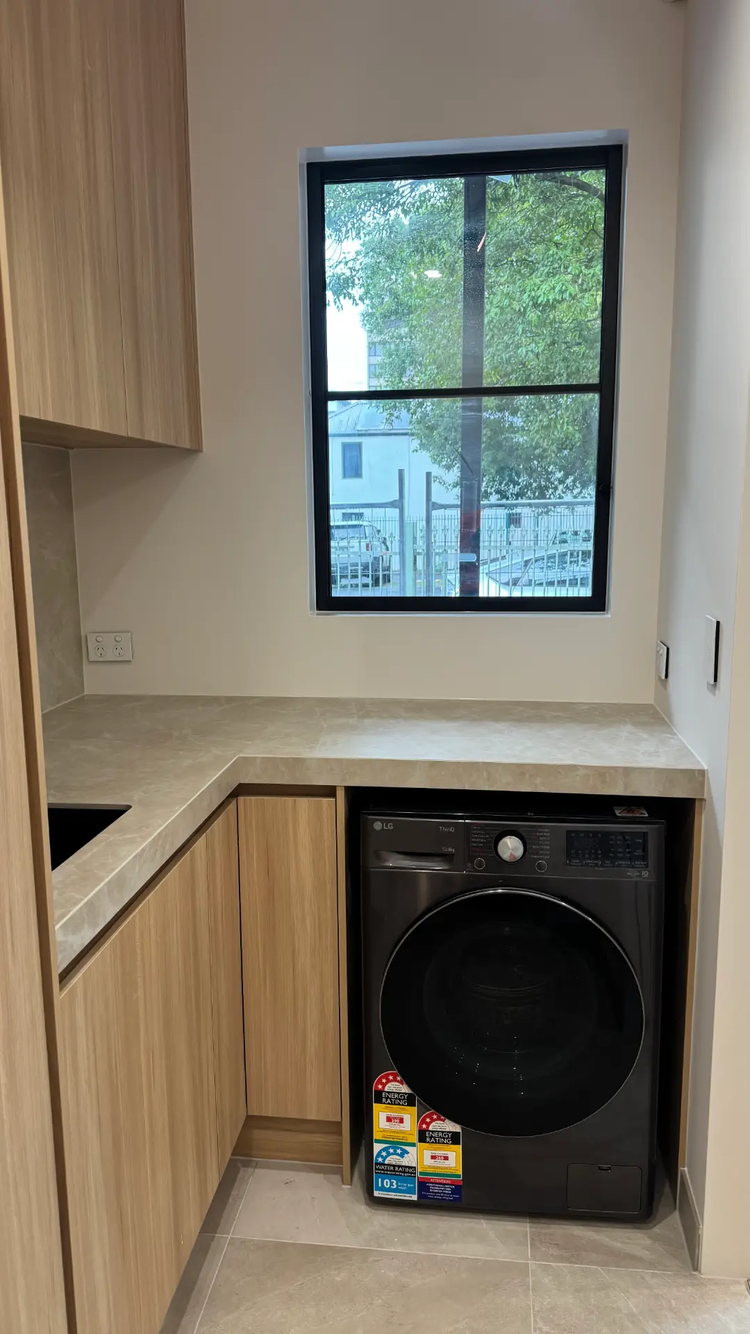 Lavandería con muebles de madera clara, encimera de piedra sellada y ventana con marco negro